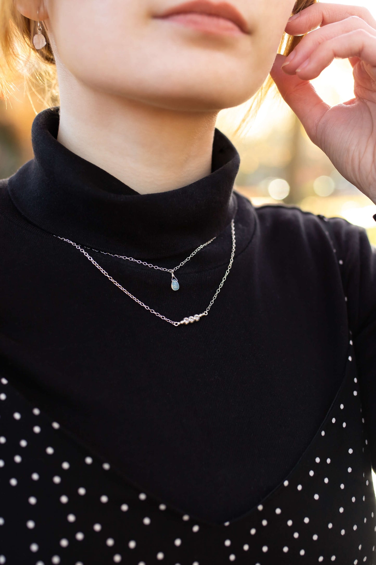 Person wearing a black turtleneck with layered necklaces, blurred outdoor background