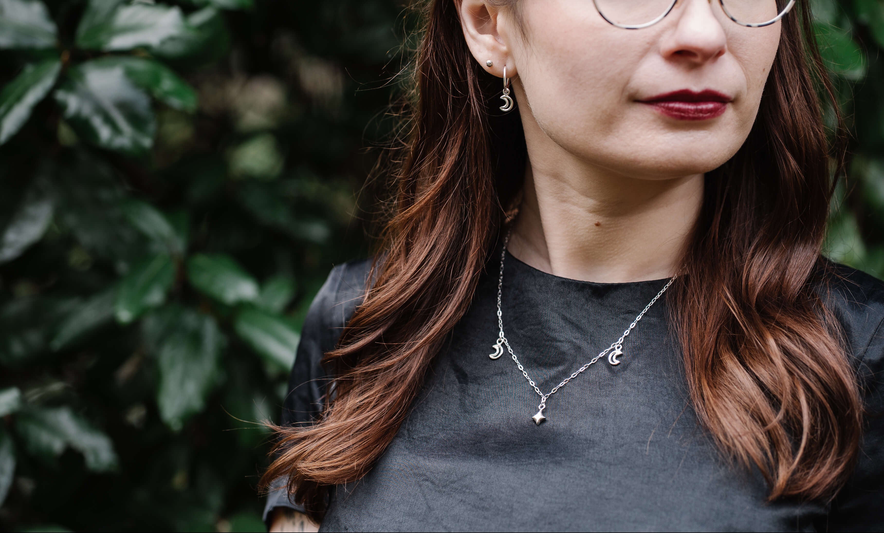 Woman wearing glasses, silver jewellery and a dark shirt with a green leafy background