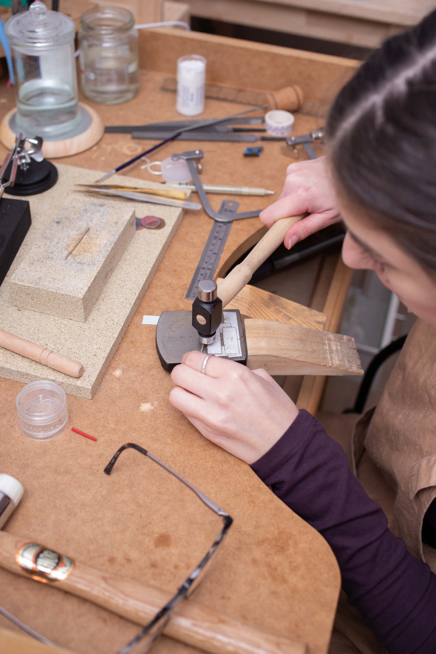 View of a jewellery bench from above with files and tools. A woman hammers a piece of silver.
